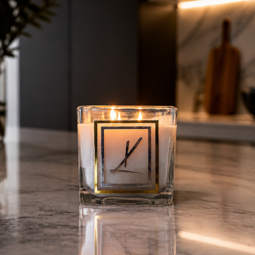 A lit double-wick candle in a clear square glass jar with a Karys Layne logo on the front, set on a polished marble countertop in a modern kitchen environment.