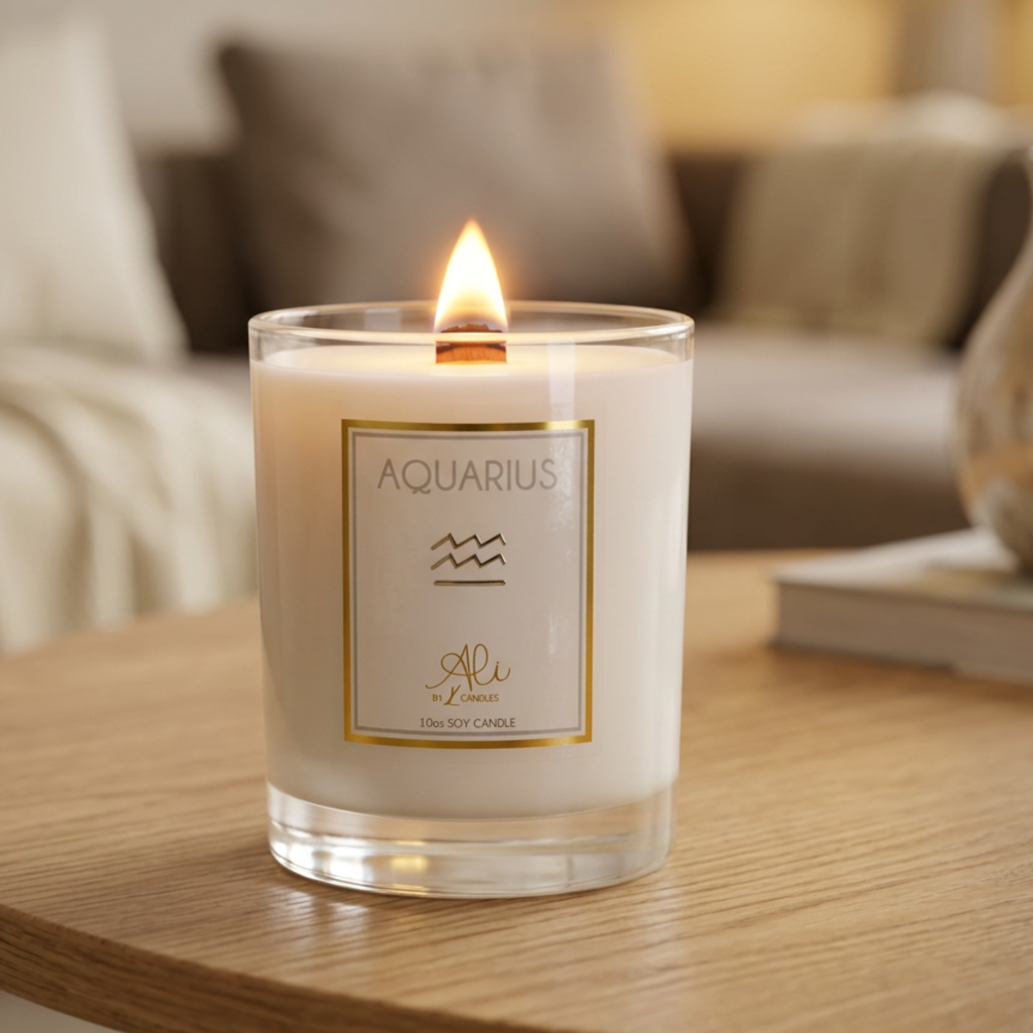 A lit soy candle in a clear glass jar sits on a wooden table in a cozy living room setting. The white wax candle features a square cream label with a gold border, the word 'AQUARIUS' at the top, a simple wave symbol, and the brand name karys layne candles at the bottom.