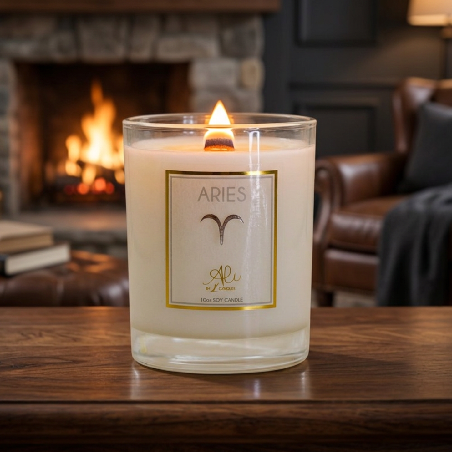 A lit Aries scented soy candle in a clear glass jar sits on a dark wooden table in front of a cozy fireplace and leather armchair. The label features the word 'ARIES" 10 oz karys layne candle
