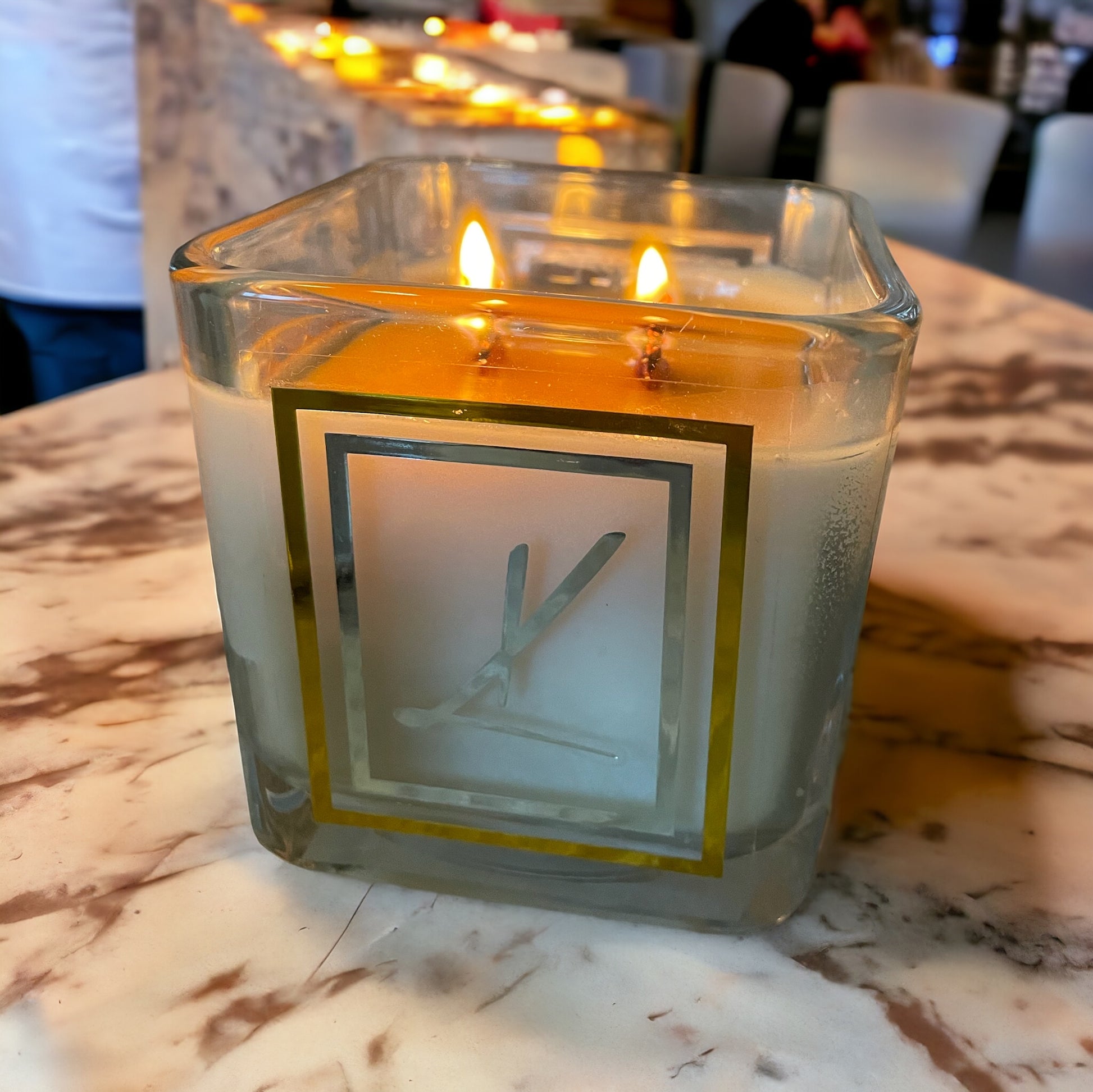 A square Karys Layne Candle with two glowing wicks sits on a cream marble countertop at a dinner party.