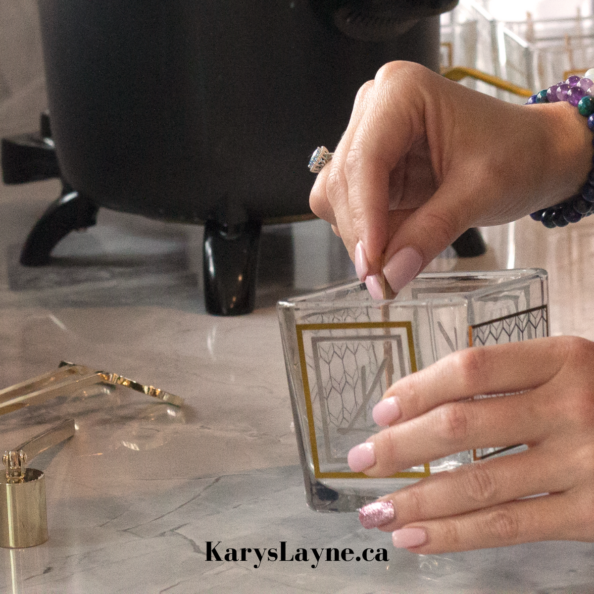 A person's hands with pink manicured nails are shown holding a clear square glass tumbler with gold geometric patterns. The background features a black tripod-like object on a marble countertop, with a gold metal accessory lying nearby.