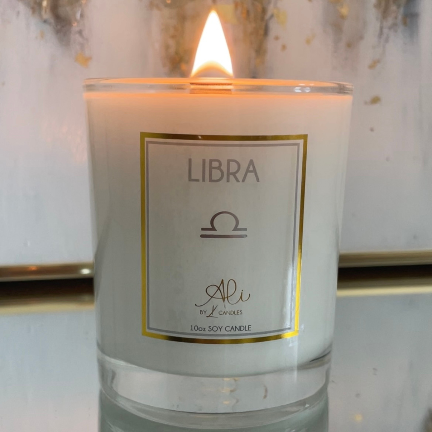 A lit 10oz soy candle in a clear glass jar with a white wax body and a white label featuring a gold border. The label reads 'LIBRA' with a Libra zodiac symbol and the brand name 'Ali by Karys Layne Candles'.