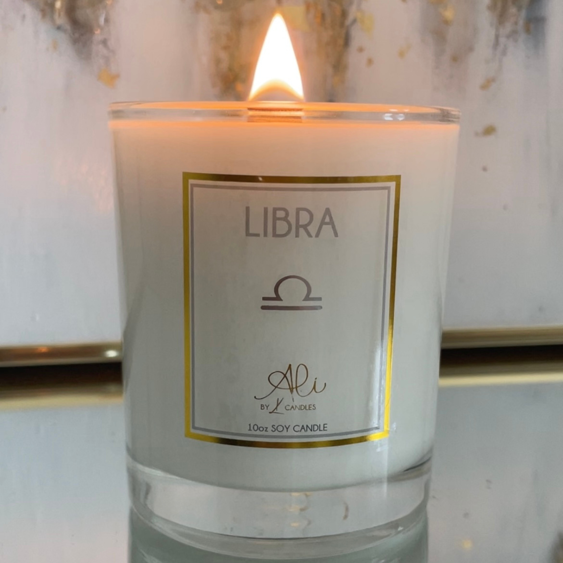 A lit 10oz soy candle in a clear glass jar with a white wax body and a white label featuring a gold border. The label reads 'LIBRA' with a Libra zodiac symbol and the brand name 'Ali by Karys Layne Candles'.