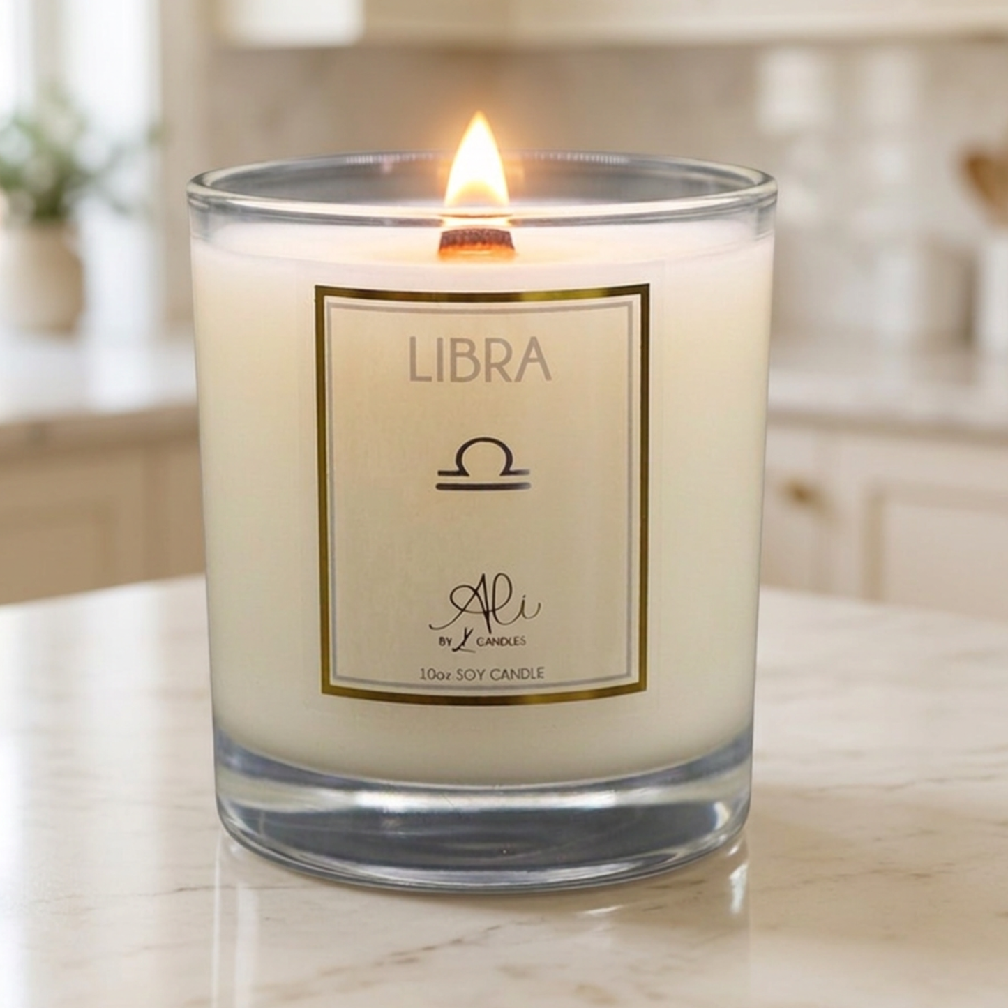 A lit soy candle in a clear glass jar sits on a light marble countertop with a softly blurred kitchen background. The front label reads 'LIBRA' with a Libra zodiac symbol and the brand name 'Ali' by Karys Layne Candles
