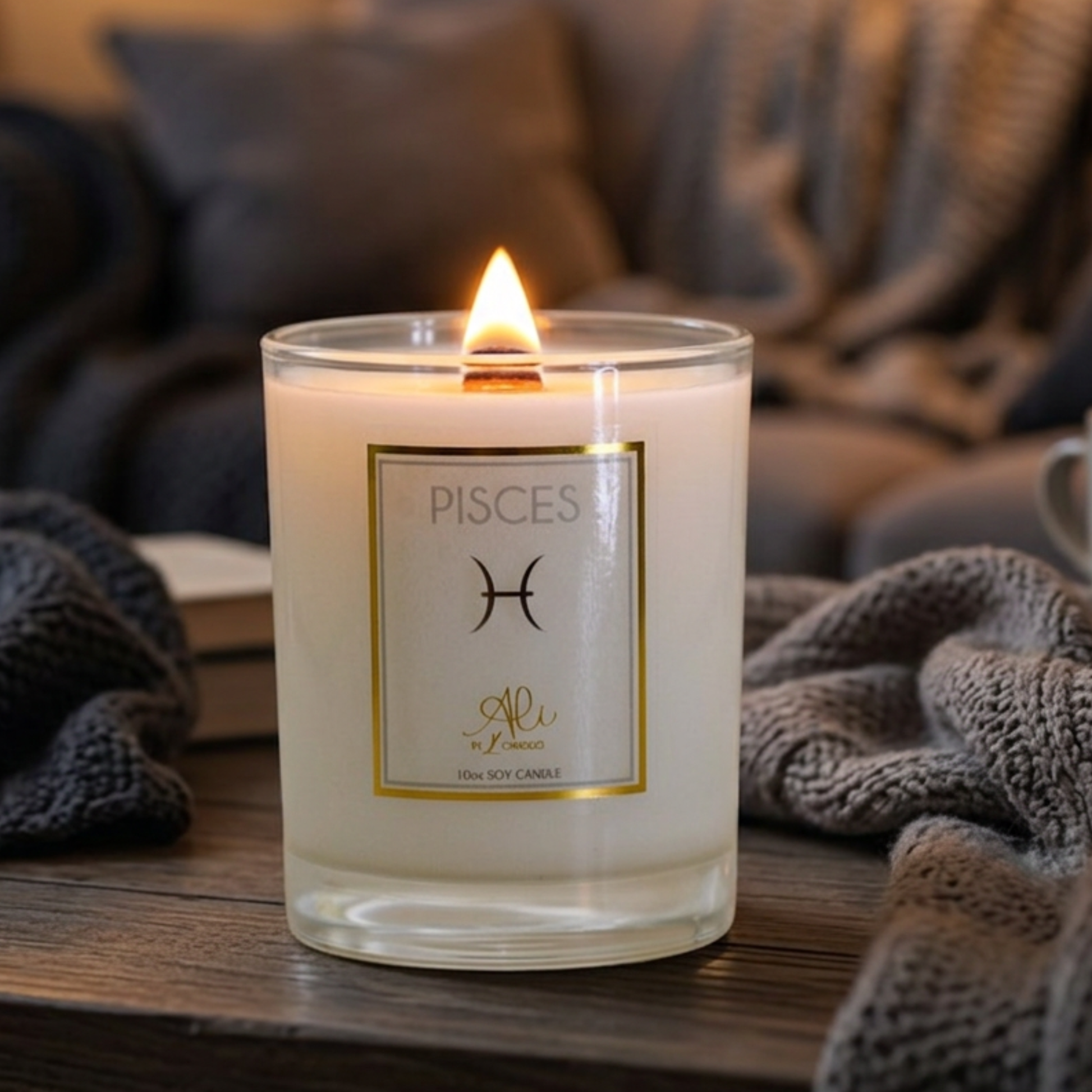 A lit white soy candle in a clear glass jar sits on a wooden table, with a cozy gray knit blanket and soft-focus home furnishings in the background. The front label reads 'PISCES' with a black astrological symbol and small gold script branding.