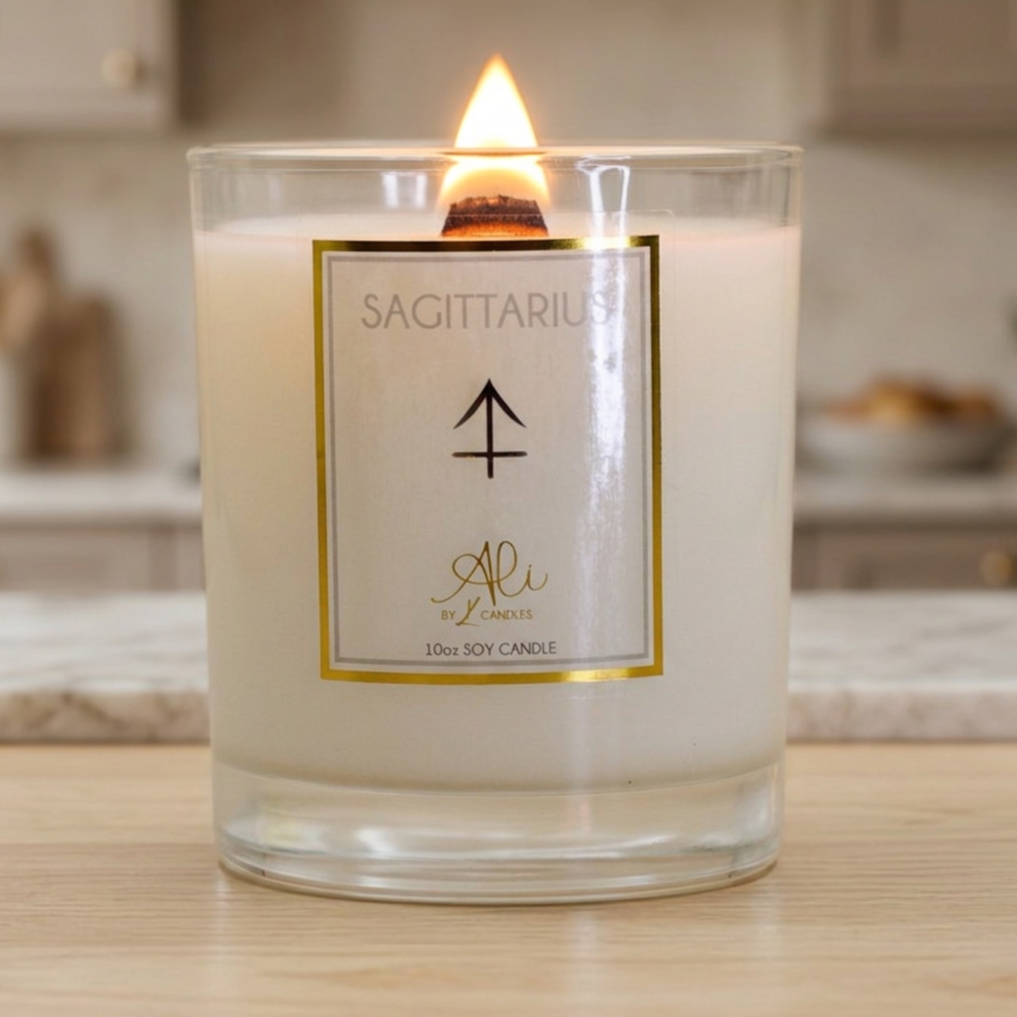 A lit 10oz soy candle in a clear glass jar with a white wax fill and a white label featuring the Sagittarius zodiac symbol and the brand name 'Ali by karys layne Candle'. The candle is set on a light wooden surface with a softly blurred kitchen background.