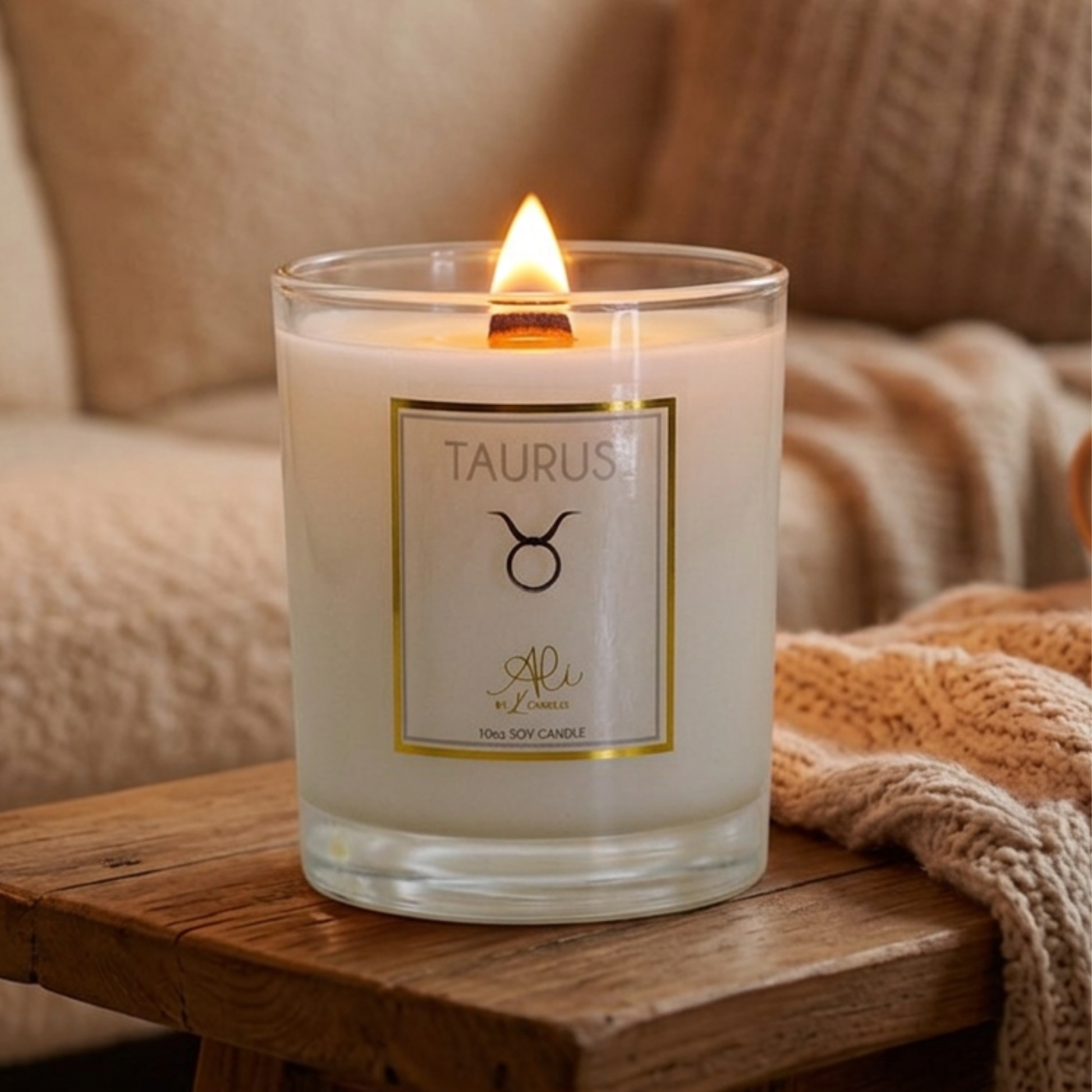 A lit 10oz soy candle in a clear glass jar sits on a wooden table in a cozy living room setting with a knitted blanket and sofa in the background. The label reads 'TAURUS' with a zodiac symbol karys layne candles