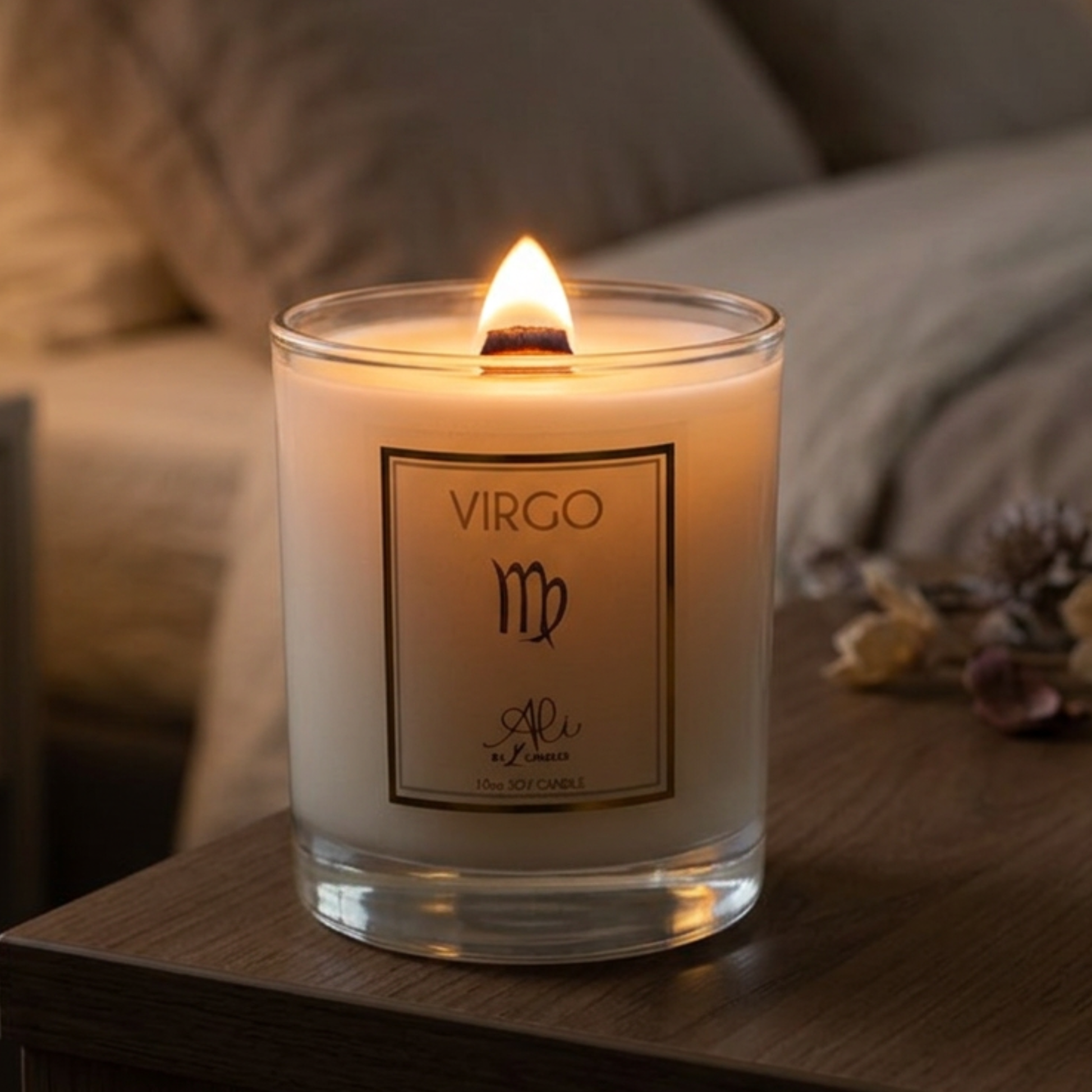 A lit white candle in a clear glass jar sits on a wooden table in a cozy bedroom setting. The front label reads 'VIRGO' with a decorative monogram and the brand name Karys Layne candles'.