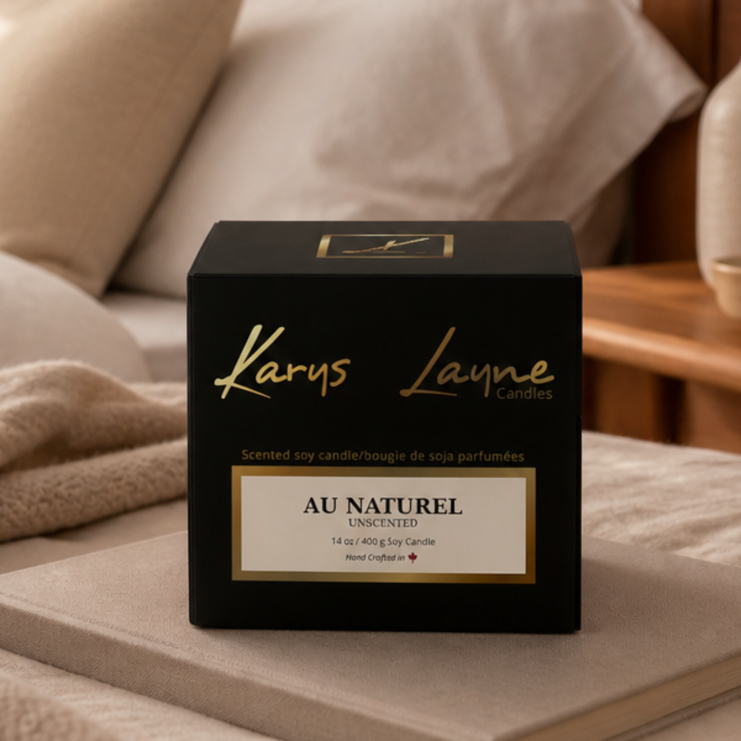 A black square box of Karys Layne Candle, scent 'Au Naturel' unscented, placed on a beige blanket in a cozy bedroom setting.