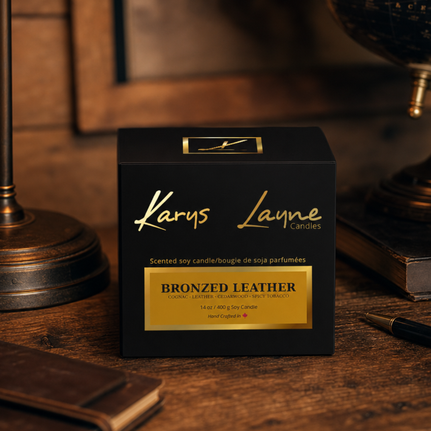 A black square candle box with gold lettering reading 'Karus Layne Candles' and a gold label that says 'Bronzed Leather,' placed on a wooden table with a lamp, books, and a pen in a warm indoor setting.