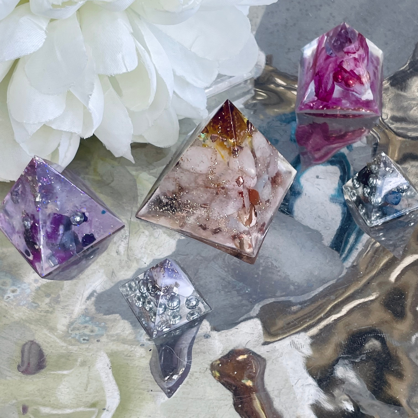 Several organite pyramids with embedded gemstones and metallic flakes are arranged on a reflective surface with a white flower in the background. The pyramids show colors including purple, pink, peach, silver, and gold.