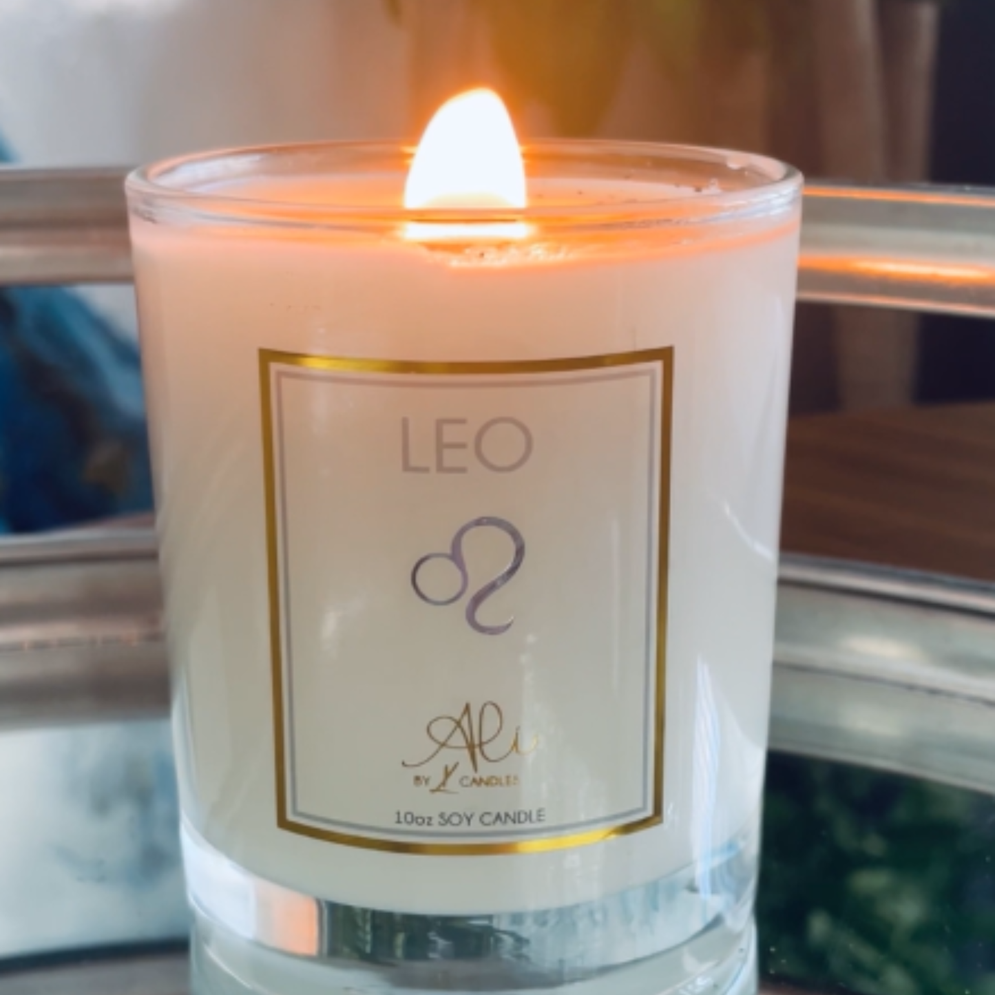 A lit white soy candle in a clear glass jar with a gold-bordered label reading 'LEO' and a purple astrological symbol, photographed close up with a blurred indoor background.