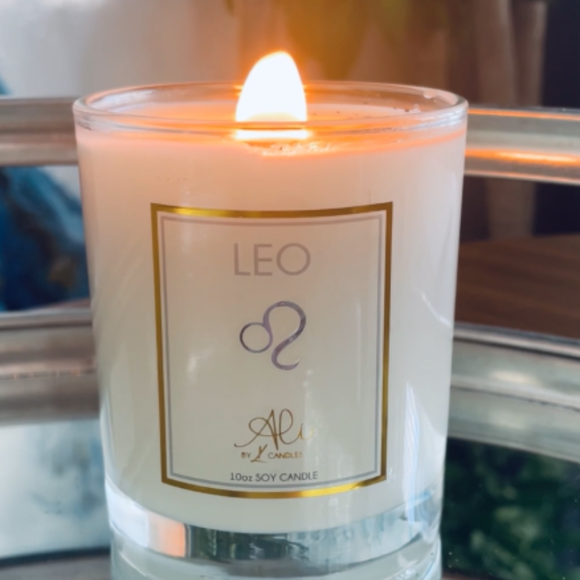 A lit white soy candle in a clear glass jar with a gold-bordered label reading 'LEO' and a purple astrological symbol, photographed close up with a blurred indoor background.