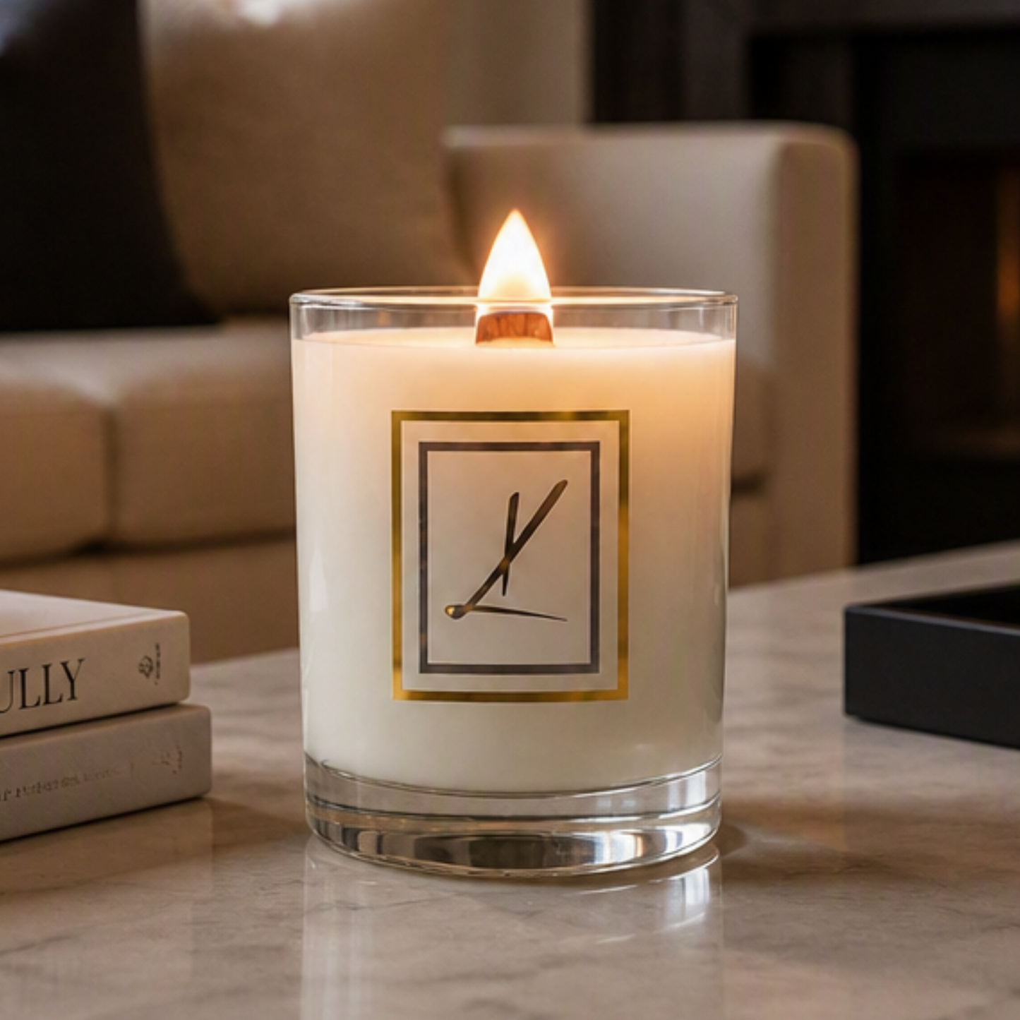 A lit white candle in a clear glass jar with a Karys Layne logo on the front, set on a marble tabletop in a cozy living room environment. Choose your scent and transform your story.