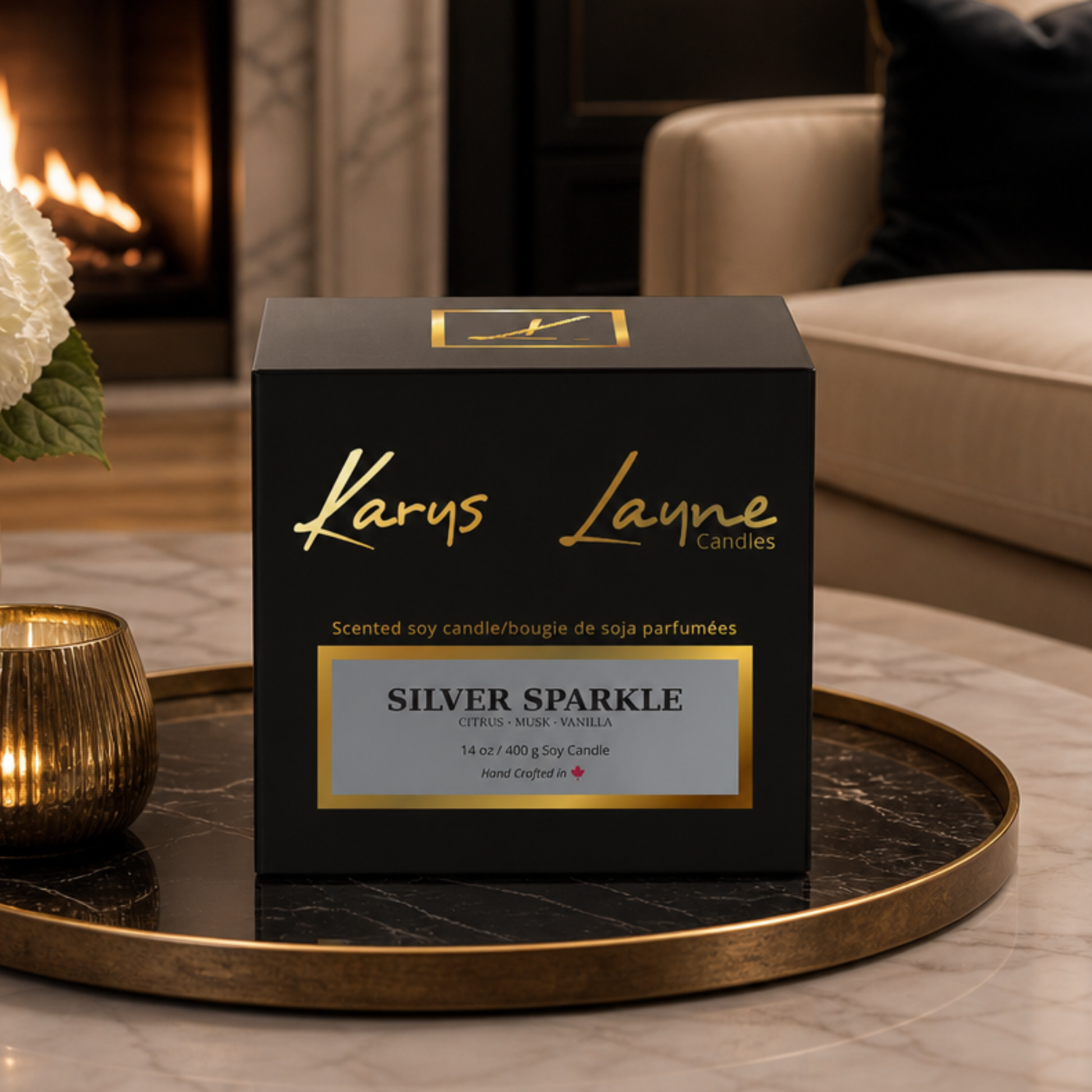 A black Karys Layne Candles box labeled Silver Sparkle, placed on a round gold tray in a softly lit living room with a fireplace and sofa in the background.
